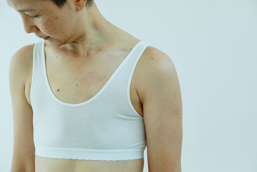 Crop ethnic female with short hair in white bra and operated breast standing against white background and looking down thoughtfully